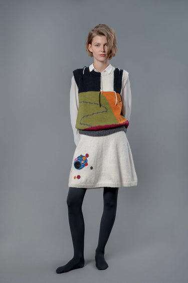 Woman wearing white wool knee length skirt and wool vest. The skirt has colourful bubble-like embroideries on the right leg.
